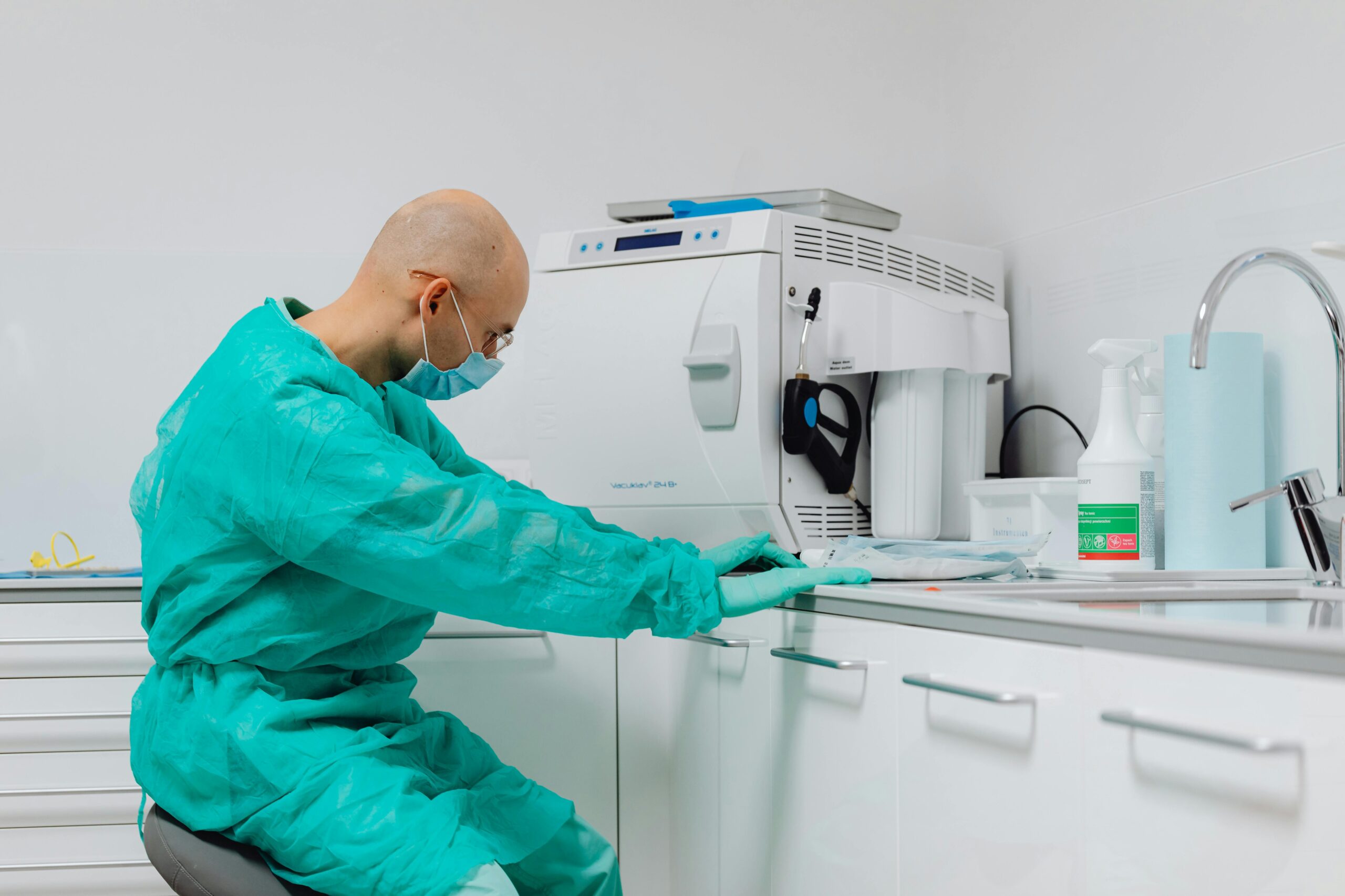 A healthcare professional in a turquoise protective gown and mask sits in a clinical setting, emphasizing the importance of precision as they work at a counter with medical supplies. A sterilization machine is visible in the background, highlighting the critical role of hygiene in diagnostic lab tests and dentistry.