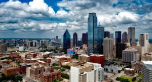 Aerial view of a city skyline with tall skyscrapers under a partly cloudy sky. Red and white buildings are visible in the foreground, with a mix of modern and older architecture throughout the cityscape.