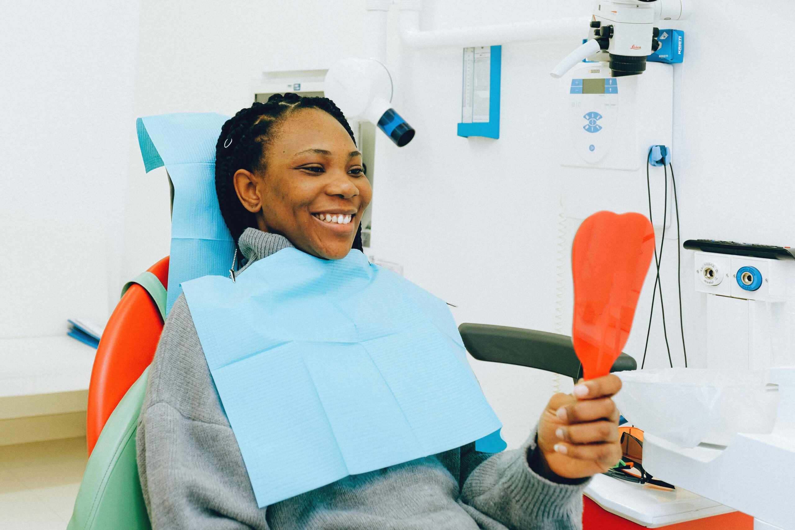 A person sits in a dental chair, smiling and holding a red mirror. Wearing a dental bib, they are at a clinic with equipment visible in the background, celebrating their oral health.